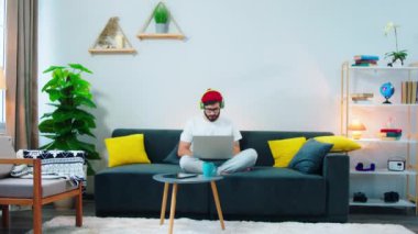 Young man with a red hat at home in the living room on the sofa using laptop to work online he concentrated typing some documents and listening the music from the headphones at the same time