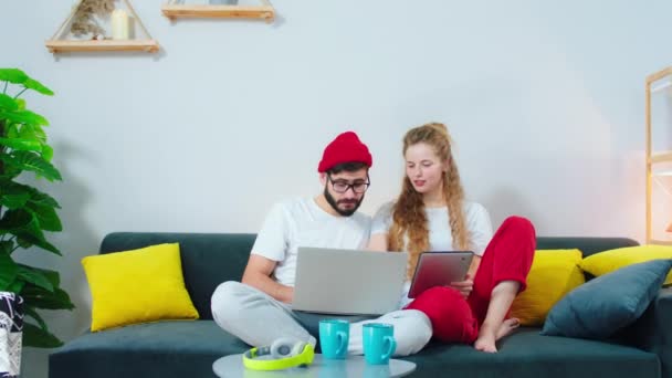 Nel soggiorno sul divano bella signora capelli biondi e il suo ragazzo trascorrere un buon tempo insieme a guardare sul computer portatile e tablet qualche bel film e avere una buona discussione — Video Stock