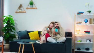 A young couple shivering on the couch and drinking something out of a blue mug, looking directly at the camera