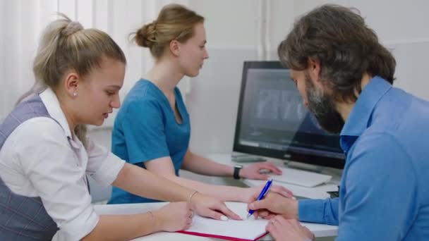 L'uomo maturo paziente firma prima di iniziare il controllo dello stato di salute dell'assistente della clinica dando alcune pagine che il medico femminile fa alcuni appunti sul computer. Girato su ARRI Alexa — Video Stock