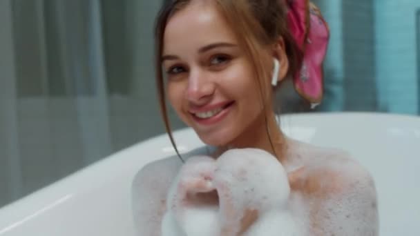 Charismatique jeune femme gros plan à la caméra souriant grand et profiter du temps dans sa salle de bain pleine de bain à bulles et écouter de la musique des écouteurs. 4k — Video