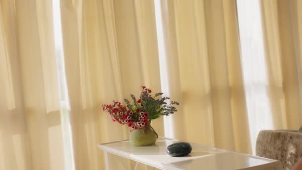 Ein Raum mit cremefarbener Palette und langen Vorhängen hat einen Tisch mit einer Vase gefüllt mit Blumen aller Art und einem Redner, eine Frau geht vorbei und schaut aus dem Fenster — Stockvideo