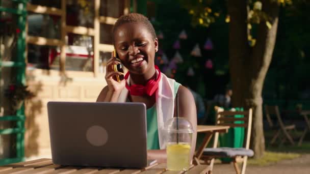 아주 행복하고 매력적 인 한 흑인 여성 이 그녀의 얼굴에 햇살이 내리쬐는 공원에 있습니다. 그녀는 테이블에 앉아 노트북에서 일을 하고 있고 그녀 옆에 레모네이드 음료가 있습니다. — 비디오