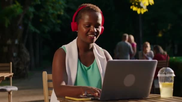 Een mooie zwarte vrouw met kort haar met een rode draadloze hoofdtelefoon sms 't op haar telefoon glimlachend en zittend aan een tafel in een park, vibing met de muziek — Stockvideo