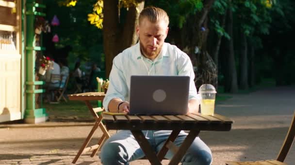 En stilig man med skägg och slickat bakhår sitter vid ett bord i parken och gör ett seriöst arbete medan han lyssnar på musik på sina hörlurar. — Stockvideo