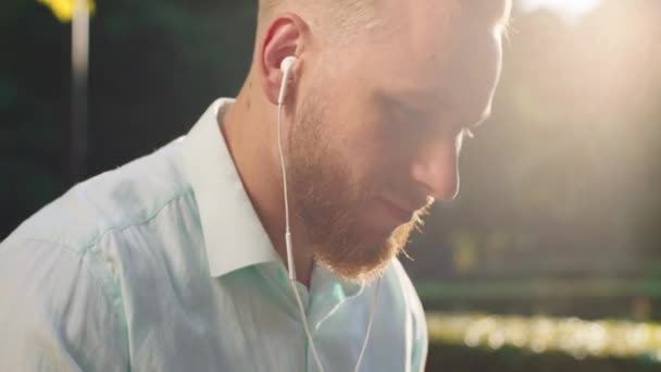 En närbild av en mycket sexig man med skägg, hans hår är blont och halkade tillbaka, han lyssnar på musik på sina hörlurar och ler medan han är utanför, också bär en grön teal — Stockvideo
