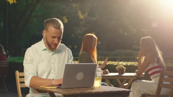 Seorang pria muda yang sangat mencolok sedang duduk di meja di kebun umum, dia sedang mengerjakan sebuah laptop dan terlihat sangat bahagia, di latar belakang dua gadis cantik sedang melakukan percakapan yang mendalam — Stok Video