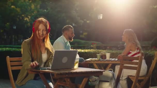 Una donna splendida con lunghi capelli rossi sani si toglie le cuffie e navigando sul suo computer portatile nel parco, prende il telefono dal tavolo. — Video Stock