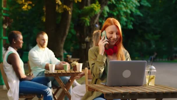 Obdivuhodná žena s dlouhými zrzavými vlasy je u stolu v blízkosti kavárny v parku, ona je v hluboké konverzaci na svém telefonu vypadá velmi šťastně, zatímco dělá pohyby rukou, vzadu skupina přátel jsou — Stock video