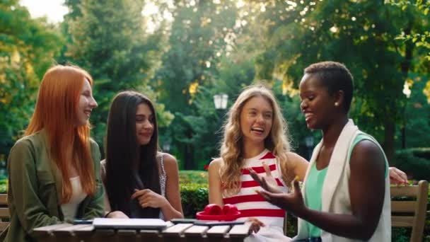 Bir grup çekici kadın parkta bir kafe masasındalar ve güzel renkli bir kadın hepsini güldüren ve güldüren bir şaka yapıyor. 4k — Stok video