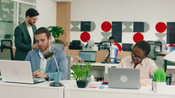In einem Büro fragt eine schöne schwarze Frau mit kurzen Haaren ihren Kollegen nach der Arbeit an ihrem Laptop, der gutaussehende Mann spricht zurück zu ihr, während sie ein Gespräch führen. — Stockvideo