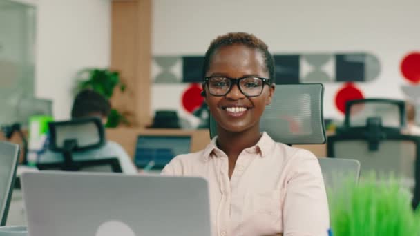 Eine intelligent aussehende Afrikanerin mit kurzen Haaren und Brille sitzt an ihrem Schreibtisch und sieht sehr fröhlich aus — Stockvideo