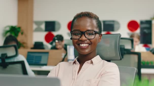 한 젊은 흑인 여성 이 카메라를 보고 있습니다. 그녀는 사무실에서 매우 행복해 보이죠. — 비디오