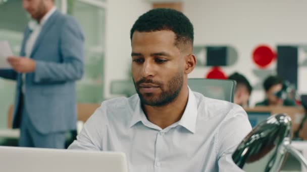 Um negro muito atraente está olhando confiante no escritório enquanto ele está fazendo seu trabalho na mesa, ele olha para cima e sorri — Vídeo de Stock