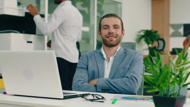 Um homem com uma barba muito atraente em um terno azul e elegante está sentado em sua mesa em um escritório estético e olhando diretamente para a câmera, tendo seus braços cruzados sorrindo para si mesmo — Vídeo de Stock