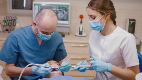Γυναίκα ασθενής έχει μια επίσκεψη στην οδοντιατρική κλινική οδοντίατρος άνθρωπος και η βοηθός του κυρία θεραπεία της τερηδόνας του ασθενούς που φορούν προστατευτική μάσκα — Αρχείο Βίντεο