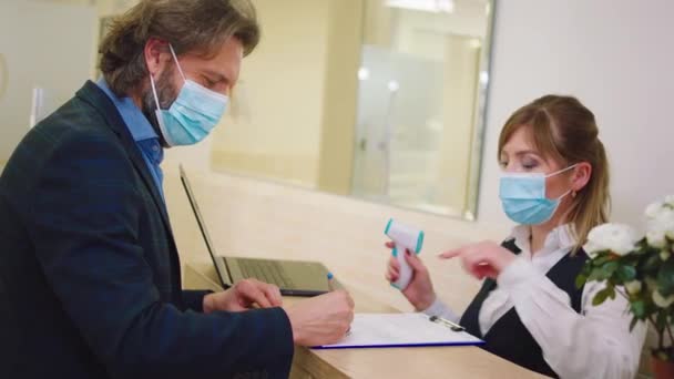 Bij de receptie van de kliniek controleert de vrouwelijke assistent de temperatuur van de patiënt man in de pandemie van het coronavirus de patiënt met het beschermende masker — Stockvideo