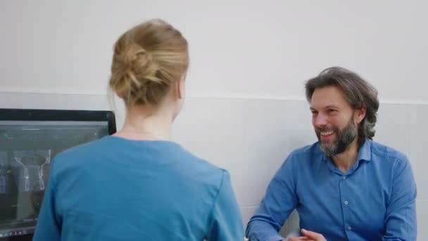W klinice dentystycznej pani dentysta i charyzmatyczny uśmiech duży pacjent rozmawia o wynikach badania skanowania patrząc na komputerze. Strzał na ARRI Alexa — Wideo stockowe