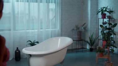 Büyüyen bitkilerle dolu büyük bir banyoda kadın banyo yapmak istiyor pembe pijamasını çıkarıyor. ARRI Alexa Mini 'de çekilen.