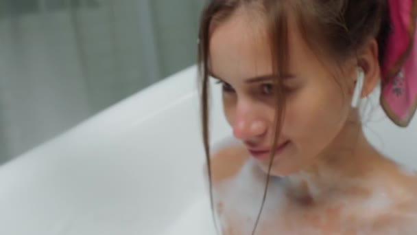 Smiling large young woman in the bathroom take a bath and enjoy the evening and listen music from the earphones — Stock Video