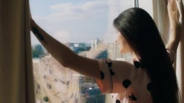 Büyüleyici bir kadın doğal perdeleri yavaşça açar ve mutlu bir şekilde gülümserken şehrin güzelliğine hayranlıkla bakar. 4k