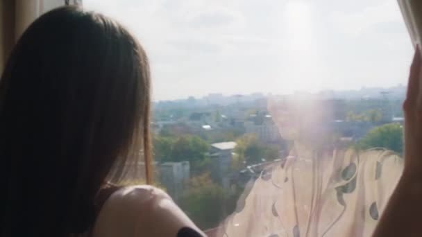 En ung kvinna öppnar spännande gardinerna för att titta ut genom fönstret mot den fantastiska utsikten över hela staden. Skjuten på ARRI Alexa Mini. — Stockvideo