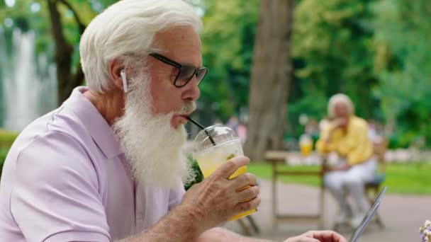 Proche de la caméra attrayant vieil homme avec des lunettes de soleil à l'aide d'un ordinateur portable pour travailler au milieu du parc au café, il dactylographier quelque chose et boire de la limonade. Tourné sur ARRI Alexa Mini. — Video