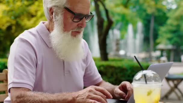 In het midden van het park in café voor de camera, oude man die op zijn laptop werkte, met een zonnebril op, concentreerde hij zich op iets typen. — Stockvideo