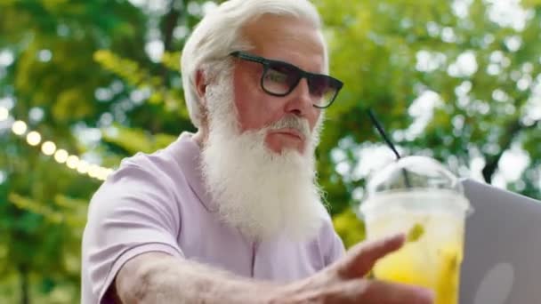 Charismatischer und gut aussehender alter Mann, der konzentriert am Laptop in der Mitte des Parks vor dem Café arbeitet, in dem er eine Sonnenbrille trägt. Schuss auf ARRI Alexa Mini. — Stockvideo
