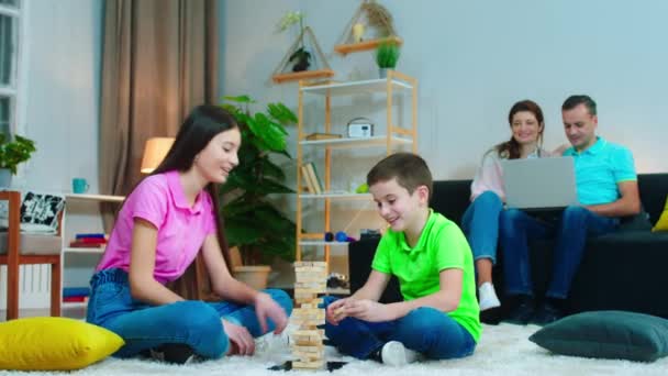 Happy boy and his big sister together playing jungle in the living room while their parents on the sofa watching some movie on the laptop — Stock Video