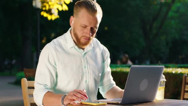 En mycket attraktiv och seriös man med skägg gör en del arbete på laptop medan du lyssnar på musik på sina hörlurar, solen strålar skiner på hans ansikte medan han är omgiven av träd — Stockvideo