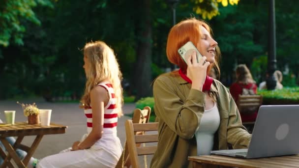 Uma mulher elegante com cabelos longos está conversando ao telefone, em um café no parque em um belo dia ensolarado, enquanto no fundo duas mulheres e um homem está sentado à mesa se divertindo e — Vídeo de Stock