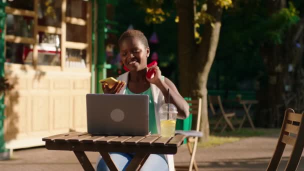 Une superbe femme noire est assise à la table seule dans un parc, elle parle au téléphone et est très joyeuse car elle fait également du travail sur son ordinateur portable. — Video