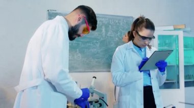 Birlikte çalışan biyoteknoloji laboratuvarı eczacıları kimyasal maddeyi cam tüpün içine karıştırıp sonuçları dijital tablete yazıyorlar.