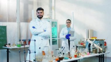 İki genç bilim adamı kimya laboratuvarında koruyucu ekipman takan çok yakışıklı bir kadın ve adam doğrudan kameraya bakıyor ve gülümsüyorlar. 4k
