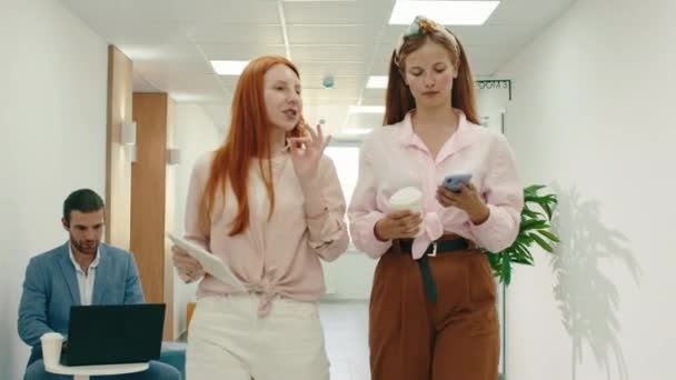 Deux belles collègues indépendantes marchent dans un couloir de bureau, la femme rousse tient une tablette tandis que la femme blonde tient son téléphone, ils parlent — Video