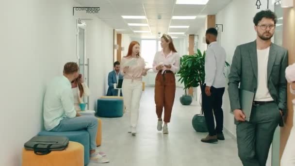 Une femme blonde et une dame aux cheveux roux sont des collègues qui se parlent en marchant dans le couloir, ils passent par beaucoup de gens, y compris un homme assis dans un sac de haricots bleus sur — Video
