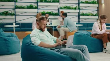 Sakallı sarışın bir adam bir oda dolusu armut ve masa başında oturmuş mavi bir fasulye torbasının üzerinde oturmuş konsantre bir şekilde bilgisayarının başında çalışıyor.