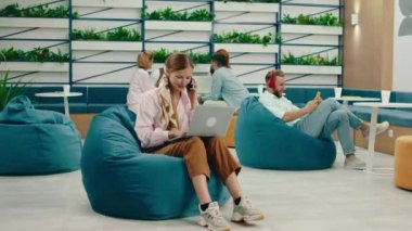 Saçında eşarp olan sarışın bir kadın laptopuyla çalışıyor ve mavi fasulye çuvalının üzerinde otururken telefonla konuşuyor.