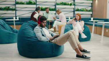 Kulaklıklarıyla müzik dinleyen yakışıklı bir adam ofis odasında içi fasulye dolu sandalyelerle oturuyor ve bilgisayarını tamir ederken arkadaki bayan telefonunu alıyor.