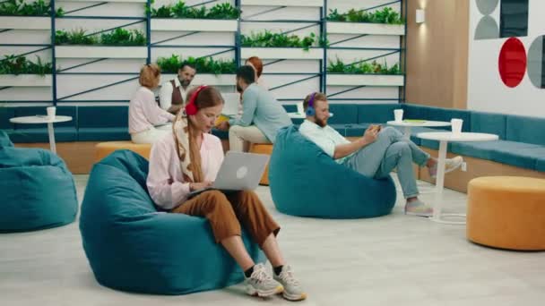 A woman is rocking out and singing with the music on her red wireless headphone while sitting on a bean bag chair doing work and in the back a group of workers are holding a meeting also in bean bag — Stock Video