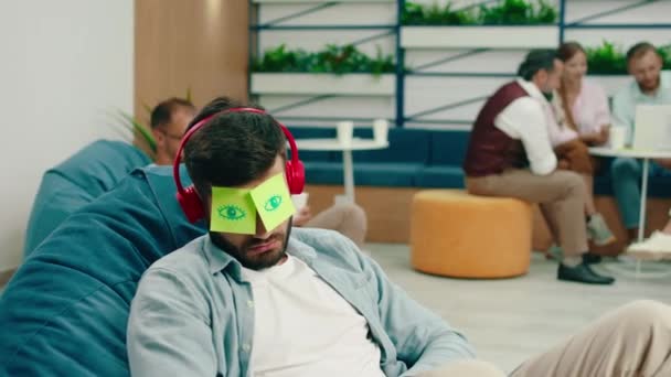 Un uomo con gli auricolari rossi senza fili sta dormendo su una sedia a sacco di fagioli e ha due appiccicosi appesi agli occhi mentre due persone dietro di lui sono sui loro telefoni — Video Stock