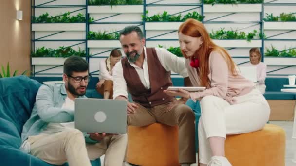 Três colegas estão tendo uma conversa interessante sobre o trabalho enquanto olham para o homem em uma cadeira de saco de feijão segurando um laptop — Vídeo de Stock