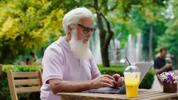 Goed uitziende oude man neemt zijn laptop mee naar het café en werkt geconcentreerd hij draagt een zonnebril — Stockvideo