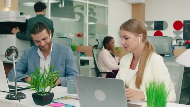 Um homem bonito branco e mulher bonita ambos vestindo ternos estão no trabalho no escritório, conversando entre si enquanto eles trabalham, no fundo uma mulher negra está falando em sua cadeira e lá — Vídeo de Stock