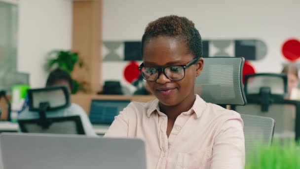 Uma jovem mulher negra muito inteligente e charmosa com óculos está em seu grande escritório, sentada à mesa e sorrindo muito — Vídeo de Stock