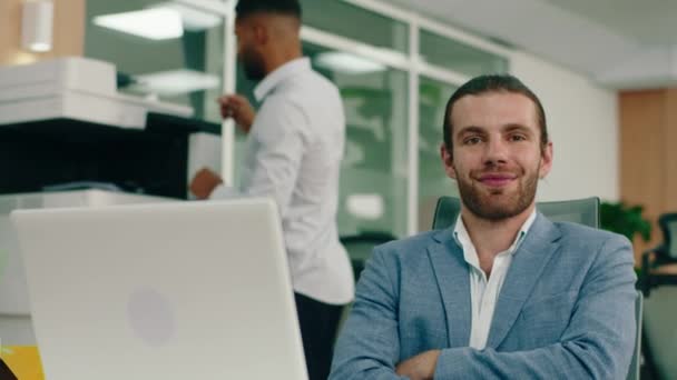 Um tiro de perto de um homem no escritório, cruzando os braços enquanto se senta em sua mesa de trabalho e sorrindo amplamente mostrando seu belo sorriso, nas costas um gerente está na impressora. 4K — Vídeo de Stock
