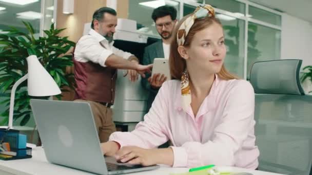 Une femme aux cheveux blonds portant un bandeau jaune est à son bureau en train de parler pendant qu'elle travaille, dans le dos deux beaux hommes de travail parlent — Video
