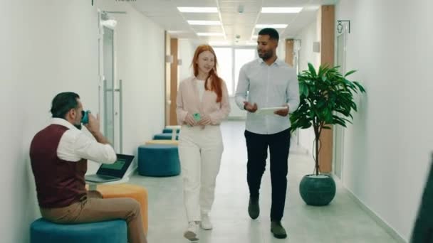 Un hombre negro está caminando con una hermosa mujer pelirroja en la oficina, se sientan en una zona de estar dentro de la oficina y detrás de ellos tres personas están haciendo trabajo en mesas de café — Vídeos de Stock