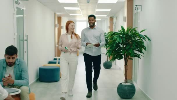 Un bel homme noir marche avec une superbe femme rousse, ce sont des collègues de travail et l'homme noir tient une tablette parlant du travail avec la femme, ils sont assis dans un bureau moderne — Video